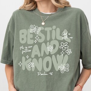 May include: Sage green t-shirt with the words "BE STILL AND KNOW" in a whimsical font, along with the phrases "He is our fortress" and "God is with us." The design includes butterfly and flower illustrations. Psalm 46 is also printed on the shirt.