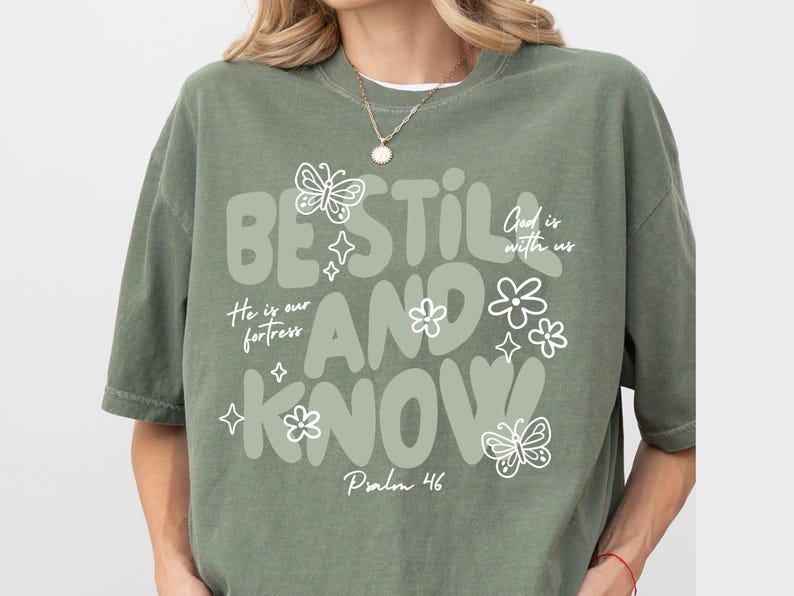 May include: Sage green t-shirt with the words "BE STILL AND KNOW" in a whimsical font, along with the phrases "He is our fortress" and "God is with us." The design includes butterfly and flower illustrations. Psalm 46 is also printed on the shirt.