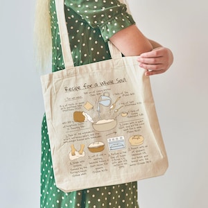May include: Cream-colored tote bag with the text "Recipe for a Whole Soul" and a recipe illustration. The recipe includes ingredients like faith, joy, and grace. The bag is held by a person wearing a green polka dot dress.