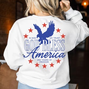 May include: A white long-sleeve sweatshirt featuring a blue eagle silhouette surrounded by red stars. The text reads: IN GOD WE TRUST, GOD BLESS AMERICA, and 250 YEARS OF FREEDOM. The design is centered on the back of the garment in a circular arrangement.