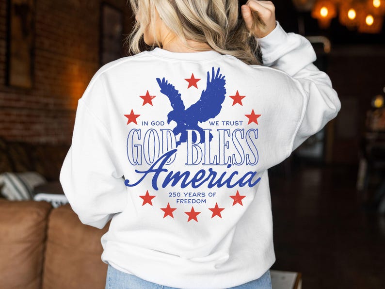 May include: A white long-sleeve sweatshirt featuring a blue eagle silhouette surrounded by red stars. The text reads: IN GOD WE TRUST, GOD BLESS AMERICA, and 250 YEARS OF FREEDOM. The design is centered on the back of the garment in a circular arrangement.