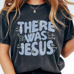 May include: Dark gray t-shirt with the text "THERE WAS JESUS" in light blue, adorned with butterfly and flower graphics. Additional text includes "in the waiting", "in the healing", "in the hurting", and "in the searching".