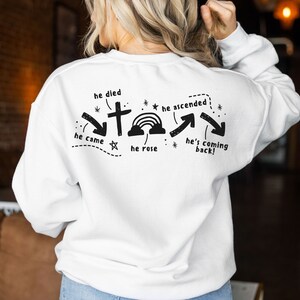 May include: White sweatshirt with a black graphic design on the back. The graphic features a cross, a rainbow, and arrows with the text "he died", "he came", "he rose", "he ascended", and "he's coming back!"