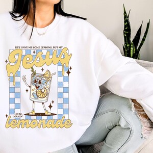 Op de afbeelding: Witte sweatshirt met een afbeelding van een cartoon limonadeglas met de tekst "LIFE GAVE ME SOME LEMONS, BUT MY JESUS MAKIN' LEMONADE". Het ontwerp heeft een blauw-wit geblokte achtergrond en gele accenten.
