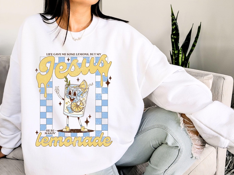 Op de afbeelding: Witte sweatshirt met een afbeelding van een cartoon limonadeglas met de tekst "LIFE GAVE ME SOME LEMONS, BUT MY JESUS MAKIN' LEMONADE". Het ontwerp heeft een blauw-wit geblokte achtergrond en gele accenten.