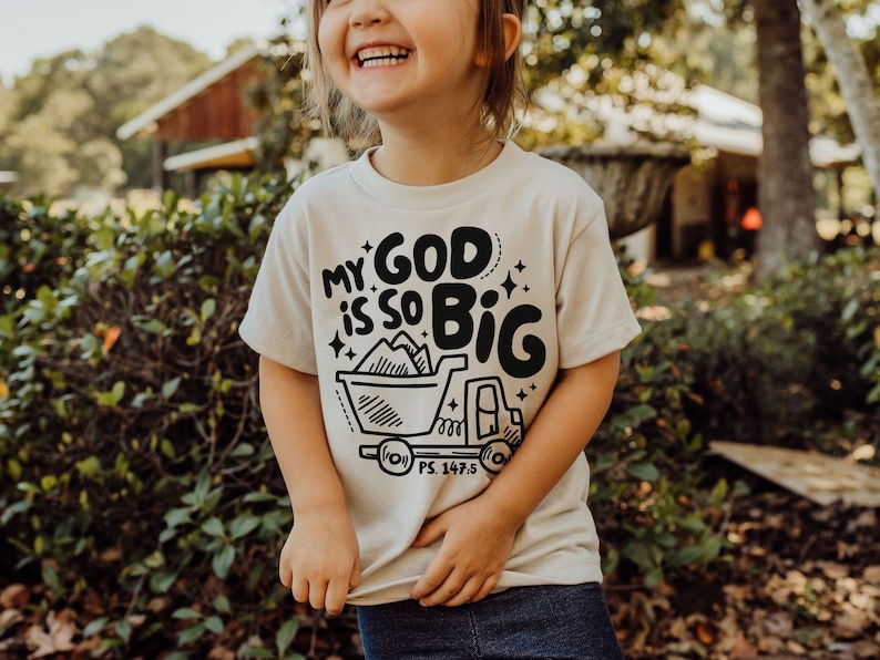May include: A child wearing a beige t-shirt with a black graphic that says "My God is so Big" and a truck with a mountain of dirt in the bed. The graphic also includes the scripture reference "PS. 147:5".