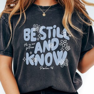 May include: Dark gray t-shirt with the words "BE STILL AND KNOW" in light blue, along with floral and butterfly graphics. The text "He is our fortress" and "Psalm 46" are also included. The shirt has a vintage, washed look.