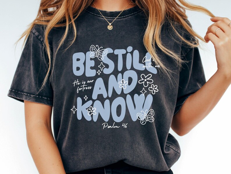 May include: Dark gray t-shirt with the words "BE STILL AND KNOW" in light blue, along with floral and butterfly graphics. The text "He is our fortress" and "Psalm 46" are also included. The shirt has a vintage, washed look.