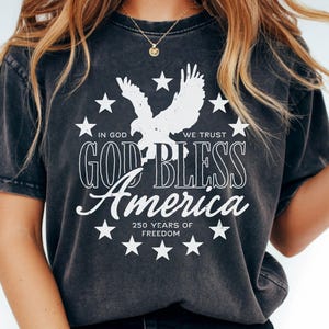 May include: A charcoal grey short-sleeve t-shirt featuring a white graphic of an eagle surrounded by stars. The text reads: IN GOD WE TRUST, GOD BLESS America, and 250 YEARS OF FREEDOM. The design is centered on the front of the garment.