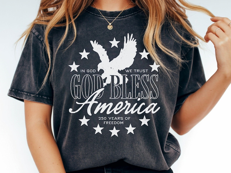 May include: A charcoal grey short-sleeve t-shirt featuring a white graphic of an eagle surrounded by stars. The text reads: IN GOD WE TRUST, GOD BLESS America, and 250 YEARS OF FREEDOM. The design is centered on the front of the garment.