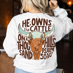 May include: White long-sleeve sweatshirt with a graphic design on the back. The design features a brown bull skull with turquoise horns, a desert landscape, and the text "He owns the cattle on a thousand hills Psalm 50:10."