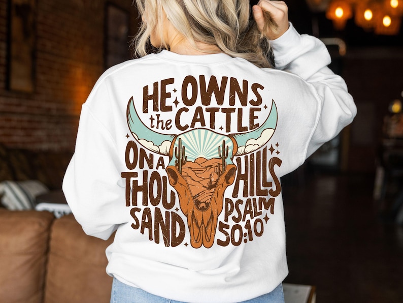 May include: White long-sleeve sweatshirt with a graphic design on the back. The design features a brown bull skull with turquoise horns, a desert landscape, and the text "He owns the cattle on a thousand hills Psalm 50:10."