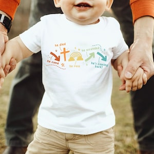 May include: A white toddler t-shirt with a colorful graphic design featuring a cross, a rainbow, and the words "he died", "he came", "he rose", "he ascended", and "he's coming back!"