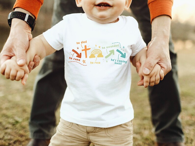 May include: A white toddler t-shirt with a colorful graphic design featuring a cross, a rainbow, and the words "he died", "he came", "he rose", "he ascended", and "he's coming back!"