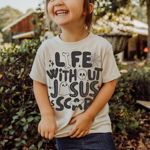 May include: A cream-colored short-sleeve t-shirt featuring dark gray text that reads Life Without Jesus Is Scary. The design includes graphic elements such as a bone, a spider web, a skull, a ghost, a small spider, stars, and a bat silhouette.