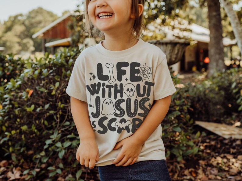 May include: A cream-colored short-sleeve t-shirt featuring dark gray text that reads Life Without Jesus Is Scary. The design includes graphic elements such as a bone, a spider web, a skull, a ghost, a small spider, stars, and a bat silhouette.