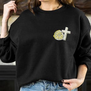 May include: Black sweatshirt with a small embroidered design featuring a white cross and a lemon slice. The lemon slice is yellow with a white rind and a cross-section visible. The sweatshirt is worn with blue jeans.