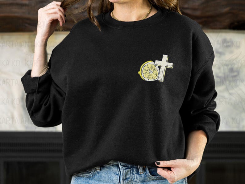 May include: Black sweatshirt with a small embroidered design featuring a white cross and a lemon slice. The lemon slice is yellow with a white rind and a cross-section visible. The sweatshirt is worn with blue jeans.