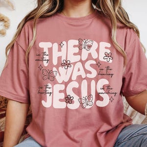 May include: Dusty rose t-shirt with the words "THERE WAS JESUS" in a retro font. The design includes butterflies and flowers. The text "in the waiting, in the hurting, in the healing, in the searching" is also included.