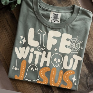 May include: A sage green t-shirt featuring the white and orange text "LIFE WITHOUT JESUS" in a bold, rounded font. The design includes graphic elements such as a spider web, a skull, a ghost, a bone, a small spider, and several four-pointed stars.