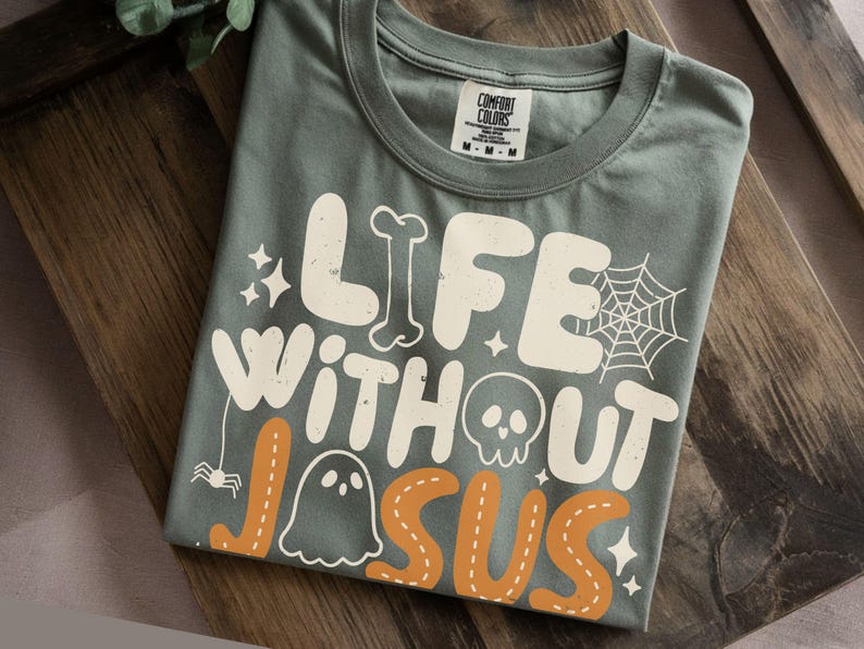 May include: A sage green t-shirt featuring the white and orange text "LIFE WITHOUT JESUS" in a bold, rounded font. The design includes graphic elements such as a spider web, a skull, a ghost, a bone, a small spider, and several four-pointed stars.