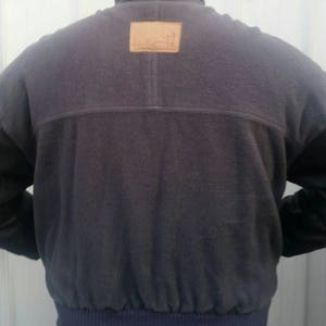 May include: A dark gray fleece jacket with a brown leather patch on the back. The jacket has a zipper closure and a ribbed waistband.