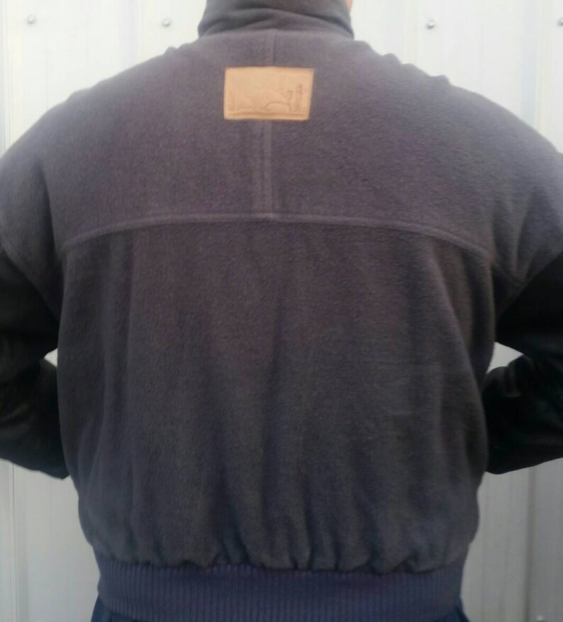May include: A dark gray fleece jacket with a brown leather patch on the back. The jacket has a zipper closure and a ribbed waistband.