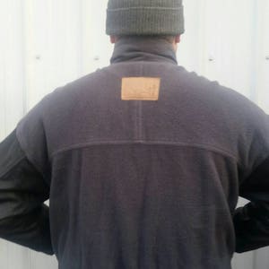 May include: A dark gray fleece jacket with a brown leather patch on the back. The jacket has a high collar and a ribbed hem. The sleeves are made of a black leather material.