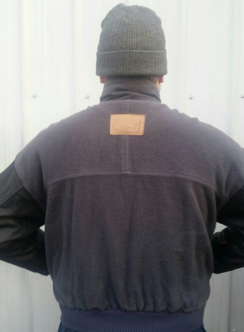May include: A dark gray fleece jacket with a brown leather patch on the back. The jacket has a high collar and a ribbed hem. The sleeves are made of a black leather material.