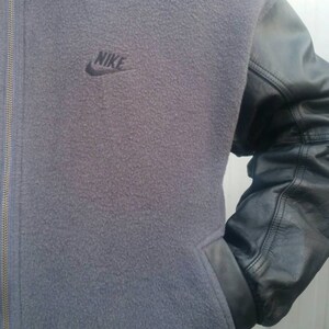 May include: A gray fleece jacket with black leather sleeves and a black Nike logo on the front. The jacket has a zipper closure.