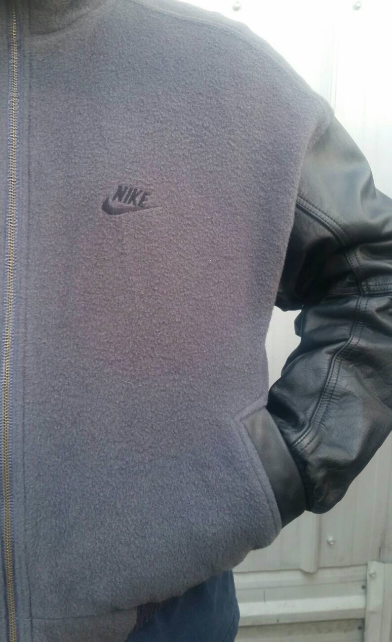 May include: A gray fleece jacket with black leather sleeves and a black Nike logo on the front. The jacket has a zipper closure.