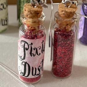 May include: Glass bottle earrings filled with red glitter, cork stoppers, and silver earring hooks. One bottle has a label that reads "Pixie Dust" in black script on a pink background. The earrings are a whimsical accessory.