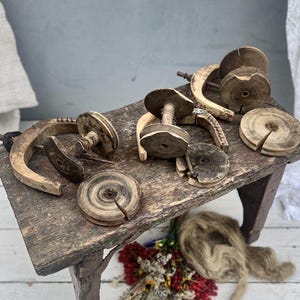 May include: A collection of weathered wooden spinning wheel parts, including several circular bobbins and curved wooden frames, arranged on a rustic, dark-stained wooden bench. The components show visible wood grain, natural wear, and aged textures.