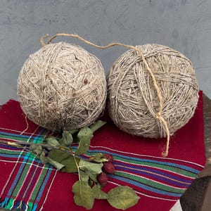 Op de afbeelding: Twee beige garenballen liggen op een kleine, rustieke houten kruk. Een rode geruite sjaal met groen, blauw en witte strepen is over de kruk gedrapeerd. Een takje met bladeren en bessen voegt een natuurlijk tintje toe.