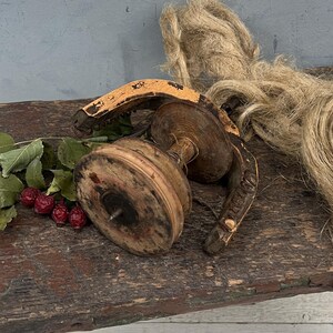 Può includere: Uno sgabello rustico in legno con finitura anticata, che tiene un oggetto in legno vintage, un fascio di fibra naturale e un rametto di bacche. Lo sgabello è dipinto in tonalità marroni e rosse.