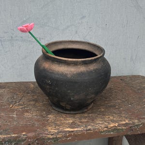 以下が含まれることがあります： 緑色の茎にピンクの花が付いた、濃い茶色の陶器の鉢。 鉢は、風化した茶色の木製のスツールの上に置かれています。 スツールは使い古された仕上がりです。 背景はプレーンなライトグレーの壁です。