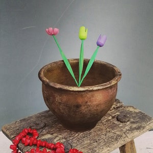 May include: A rustic wooden stool supports a bronze-colored ceramic pot with three colorful tulip-shaped flowers. A strand of red beads rests on the stool. The background is a muted gray, and the floor is white.