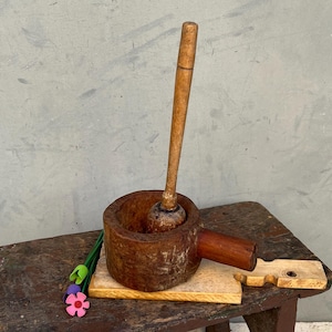 Puede incluir: Un mortero y maja de madera rústicos con una pequeña tabla de cortar. El mortero es de color marrón oscuro y la maja es de color marrón claro. El conjunto está sobre un pequeño taburete de madera desgastada. Unas flores de tela coloridas están en el taburete.