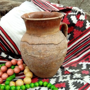 May include: A brown clay jug with a handle sits on a red, white, and black patterned fabric. Two wooden bead necklaces, one green and one pink, are on the fabric.