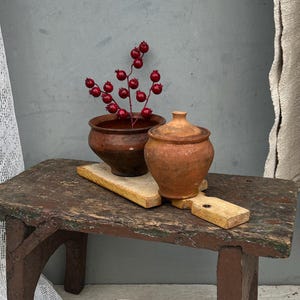 Puede incluir: Un taburete pequeño de madera marrón desgastada con cuatro patas, que sostiene una tabla de cortar de madera clara. Sobre la tabla hay un cuenco de arcilla marrón oscuro con una rama de bayas rojas y una olla de arcilla marrón claro con tapa.