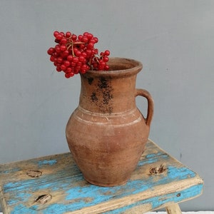 May include: A rustic brown clay pitcher with a handle sits on a blue and white painted wooden stool. A sprig of red berries is in the pitcher.