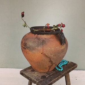 Può includere: Un vaso di terracotta rustico con una smaltatura scheggiata si trova su uno sgabello di legno. Il vaso è pieno di un ramo secco con bacche rosse. Una collana di perle turchesi è drappeggiata sullo sgabello.