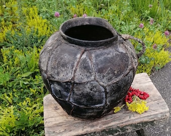 Könnte beinhalten: Ein großer, schwarzer Keramiktopf mit strukturierter Oberfläche steht auf einem Holzhocker. Der Topf hat eine breite Öffnung und einen Griff an der Seite. Der Hocker ist verwittert und hat eine grüne Lackierung.