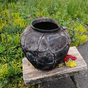 Könnte beinhalten: Ein großer, schwarzer Keramiktopf mit strukturierter Oberfläche steht auf einem Holzhocker. Der Topf hat eine breite Öffnung und einen Griff an der Seite. Der Hocker ist verwittert und hat eine grüne Lackierung.