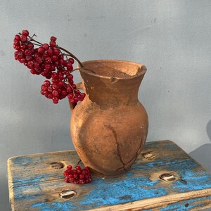 Può includere: Uno sgabello di legno rustico con vernice blu e un'anfora di terracotta con bacche rosse in cima.