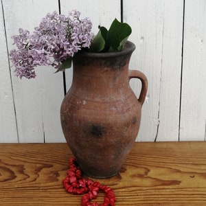 Può includere: Un vaso in terracotta con un'ampia apertura e un manico su un lato, riempito con un mazzo di fiori di lillà viola chiaro. Una collana di perle di corallo rosso si trova su un tavolo di legno davanti al vaso.