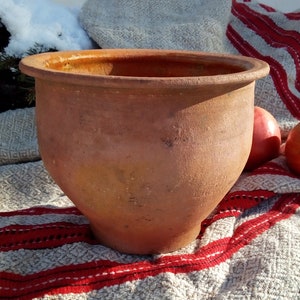 May include: A terracotta clay pot with a wide rim sits on a white and red striped blanket. The pot is slightly weathered and has a rustic appearance.