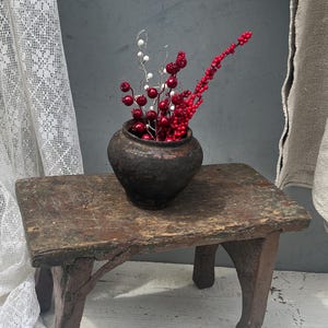 May include: A small, dark brown, weathered clay pot sits atop a low, rectangular wooden stool with four sturdy legs. Inside the pot are several thin, flexible stems adorned with clusters of small, shiny red and white decorative berries.