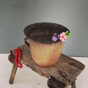 May include: A rustic wooden stool with a brown clay pot on top. The pot has a dark brown glaze and is decorated with three small plastic flowers in pink, purple, and green. A red beaded necklace is draped over the side of the stool.