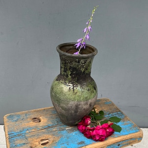 Peut inclure: Un vase en céramique vert à la texture rugueuse posé sur un tabouret en bois bleu et brun. Un brin de fleurs violettes et des roses roses sont dans le vase.
